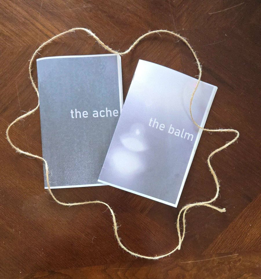 Two slightly overlapping zines with gray covers. One is titled 'The Balm,' the other is titled 'The Ache.' They are surrounded by a ring of jute twine and sit on a wooden table.